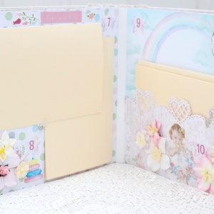 May include: A scrapbook with a light blue and pink background. The scrapbook has several pages with different designs, including a rainbow, a hot air balloon, and a baby. The scrapbook is decorated with flowers, lace, and other embellishments. The scrapbook has a number on each page, starting with 7 and ending with 10.