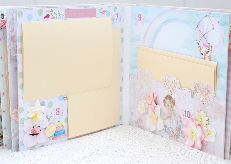 May include: A scrapbook with a light blue and pink background. The scrapbook has several pages with different designs, including a rainbow, a hot air balloon, and a baby. The scrapbook is decorated with flowers, lace, and other embellishments. The scrapbook has a number on each page, starting with 7 and ending with 10.