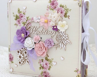 May include: A white fabric wedding album with a floral design and a wooden heart cutout with the word "Wedding" on it. The album is decorated with purple, pink, and white flowers and has a white ribbon.