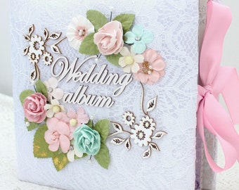 May include: White wedding album with a lace cover and a pink ribbon. The front of the album is decorated with a floral wreath made of pink, white, and blue flowers, green leaves, and wooden flowers. The words "Wedding album" are written in brown on the front of the album.