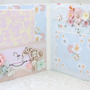 May include: A scrapbook with a light blue and pink floral pattern. The scrapbook is open to reveal two pages with a light pink and yellow patterned paper on the left page and a light blue floral patterned paper on the right page. There is a wooden cutout of a girl with a flower in her hair and a wooden cutout of a rocking horse on the left page. There is a wooden cutout of a butterfly on the right page.