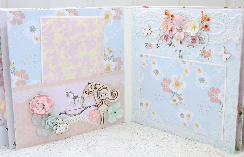 May include: A scrapbook with a light blue and pink floral pattern. The scrapbook is open to reveal two pages with a light pink and yellow patterned paper on the left page and a light blue floral patterned paper on the right page. There is a wooden cutout of a girl with a flower in her hair and a wooden cutout of a rocking horse on the left page. There is a wooden cutout of a butterfly on the right page.
