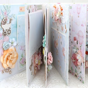May include: A scrapbook with a white background and pages decorated with floral patterns, lace, and embellishments. The pages are adorned with flowers, a baby carriage, and clouds. The scrapbook is closed and the pages are slightly open.