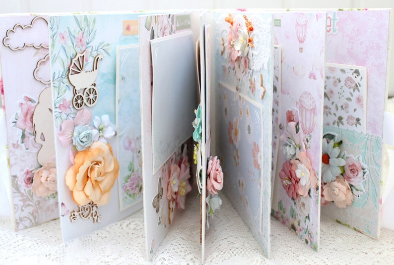 May include: A scrapbook with a white background and pages decorated with floral patterns, lace, and embellishments. The pages are adorned with flowers, a baby carriage, and clouds. The scrapbook is closed and the pages are slightly open.