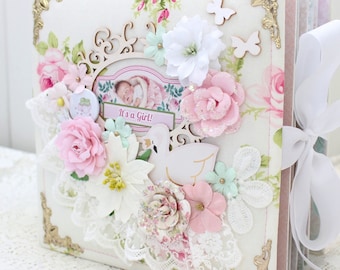 May include: A white and pink scrapbook with a floral design and a white ribbon. The cover features a wooden frame with a baby picture and the text "It's a Girl!"