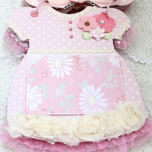 May include: Pink baby dress shaped card with a white polka dot pattern, a white lace trim, and a floral design on the front. The card has a white scalloped edge and a white ruffle trim.