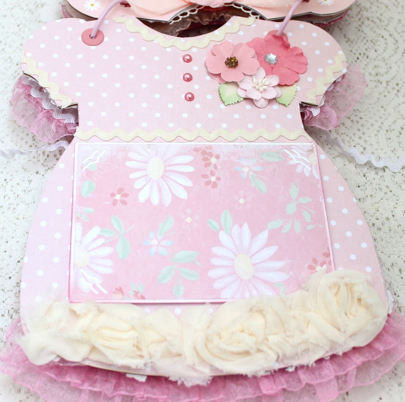 May include: Pink baby dress shaped card with a white polka dot pattern, a white lace trim, and a floral design on the front. The card has a white scalloped edge and a white ruffle trim.
