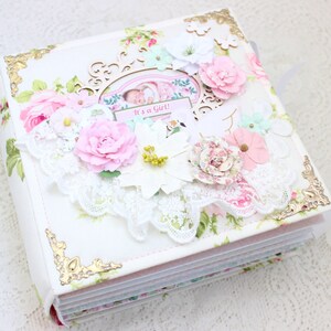 May include: A white scrapbook with a floral design and gold accents. The cover features a lace trim, flowers, and a sign that says "It's a Girl!"