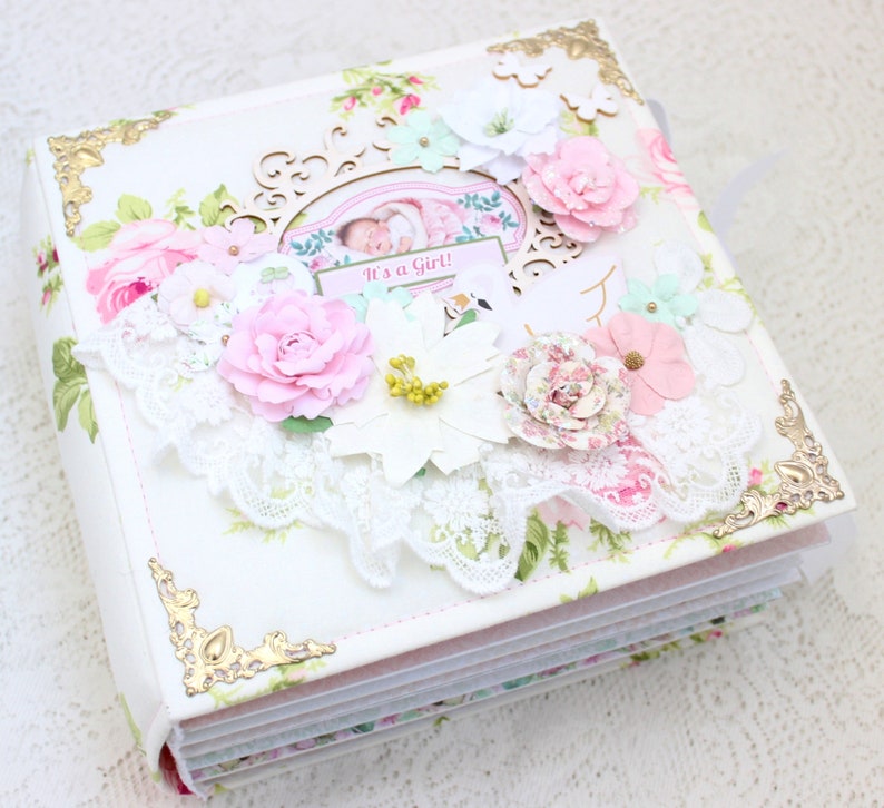 May include: A white scrapbook with a floral design and gold accents. The cover features a lace trim, flowers, and a sign that says "It's a Girl!"