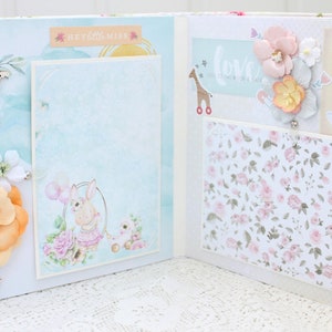 May include: A scrapbook with a light blue and white background. The scrapbook has a variety of embellishments, including flowers, a teddy bear, a baby carriage, and a giraffe. The text "Hey Little Miss" and "Love" is visible on the scrapbook.