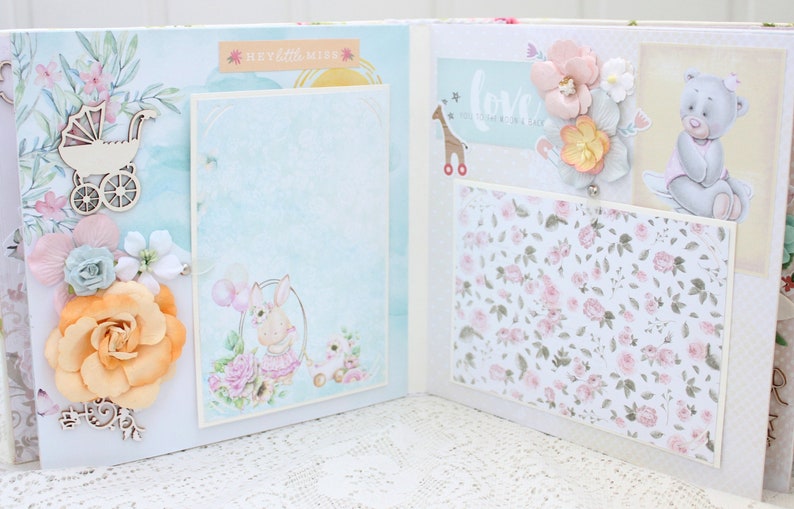 May include: A scrapbook with a light blue and white background. The scrapbook has a variety of embellishments, including flowers, a teddy bear, a baby carriage, and a giraffe. The text "Hey Little Miss" and "Love" is visible on the scrapbook.
