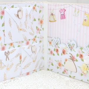 May include: A scrapbook with a floral pattern and white envelopes with wooden embellishments. The scrapbook is open to reveal a page with a pink and white striped background and a row of hanging clothes.