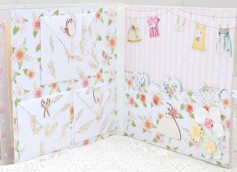 May include: A scrapbook with a floral pattern and white envelopes with wooden embellishments. The scrapbook is open to reveal a page with a pink and white striped background and a row of hanging clothes.