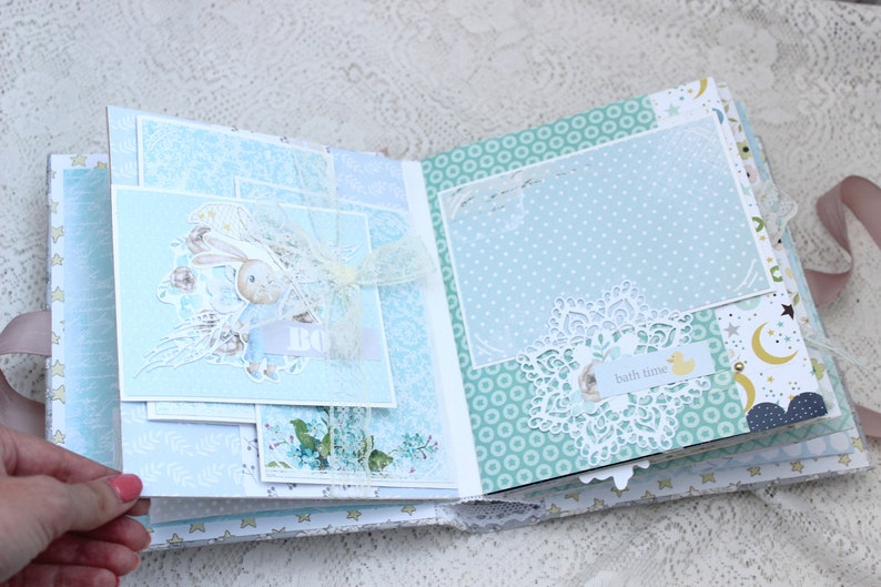 May include: A blue and white scrapbook with a bunny rabbit illustration, a white lace doily, and the text "bath time" on a yellow tag.