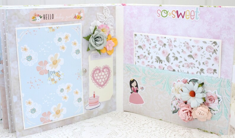 May include: A scrapbook with a light blue and white floral pattern on the left page and a pink and white floral pattern on the right page. The left page has a sticker that says "Hello" and the right page has a sticker that says "so sweet".