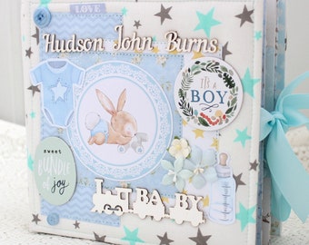 May include: A baby scrapbook with a blue and white color scheme. The cover features a wooden cutout of a train with the word "BABY" on it, a bunny rabbit illustration, and a blue and white baby onesie. The scrapbook is decorated with stars, flowers, and a ribbon. The text "Hudson John Burns" is written on the cover.