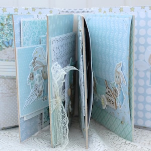 May include: A blue and white scrapbook with a bunny rabbit design. The scrapbook is decorated with lace, stars, and other embellishments. The pages are filled with photos and other mementos.
