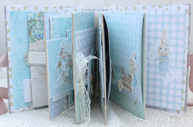 May include: A blue and white scrapbook with a bunny rabbit design. The scrapbook is decorated with lace, stars, and other embellishments. The pages are filled with photos and other mementos.