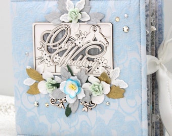 May include: A light blue wedding album with a floral pattern and the words "Our Wedding" in a decorative frame. The album is decorated with white, blue, and gold paper flowers and rhinestones. A white ribbon is tied on the side.
