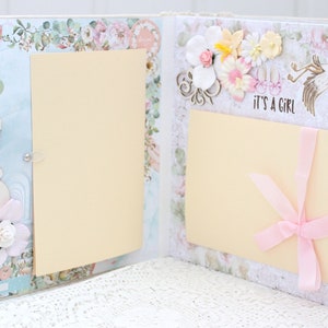 May include: A scrapbook with a light blue and white background. The scrapbook has two pages with a cream-coloured insert on each page. The left page has a cutout of a pregnant woman and flowers. The right page has a pink ribbon and the text "It's a Girl".