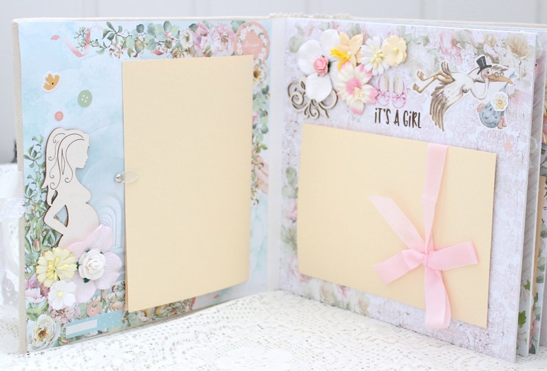 May include: A scrapbook with a light blue and white background. The scrapbook has two pages with a cream-coloured insert on each page. The left page has a cutout of a pregnant woman and flowers. The right page has a pink ribbon and the text "It's a Girl".