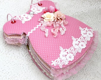 May include: A pink fabric baby dress scrapbook with white lace trim and a floral embellishment. The scrapbook is made of multiple layers of fabric and paper.