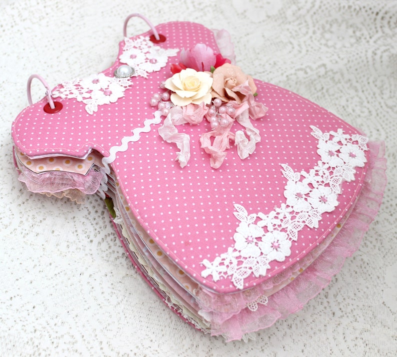 May include: A pink fabric baby dress scrapbook with white lace trim and a floral embellishment. The scrapbook is made of multiple layers of fabric and paper.