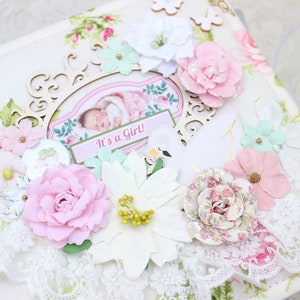 May include: A white scrapbook with a floral design and gold accents. The cover features a pink and white flower embellishment, a wooden frame with a photo of a baby, and the text "It's a Girl!"