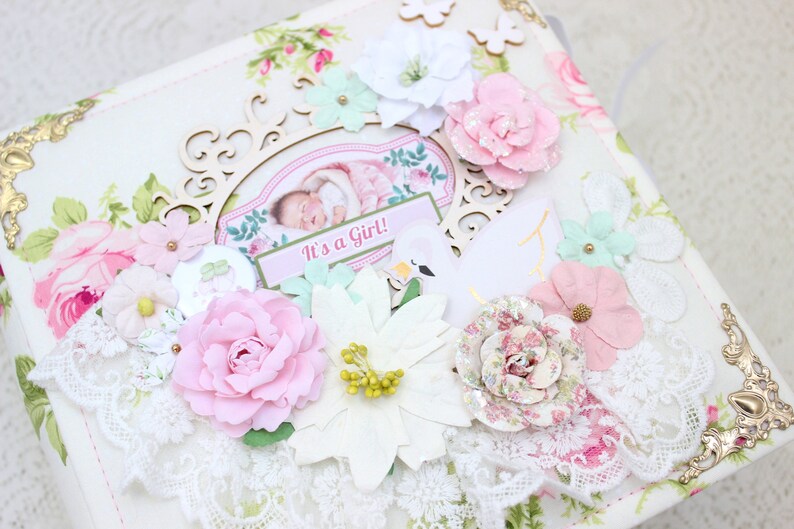 May include: A white scrapbook with a floral design and gold accents. The cover features a pink and white flower embellishment, a wooden frame with a photo of a baby, and the text "It's a Girl!"