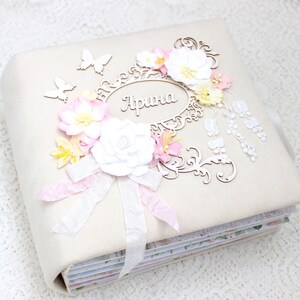 May include: A white fabric scrapbook with a decorative floral design. The scrapbook has a white ribbon and a wooden cutout with the name "Арина" in cursive.