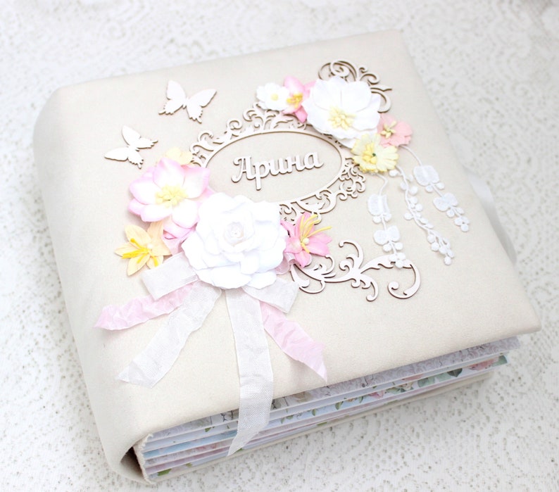 May include: A white fabric scrapbook with a decorative floral design. The scrapbook has a white ribbon and a wooden cutout with the name "Арина" in cursive.