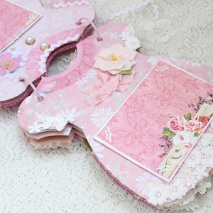 May include: Pink and white scrapbook album shaped like a dress with lace trim and floral embellishments. The album has two photo slots and a blank page for journaling.