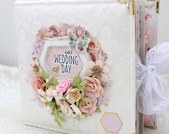 May include: White wedding album with a floral embellishment on the front cover. The embellishment features a wreath of pink and cream flowers, green leaves, and a butterfly. The album has a gold corner design and a white ribbon.