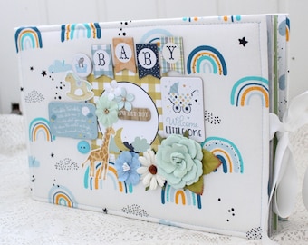 May include: A white fabric baby scrapbook with a blue, yellow, and orange rainbow pattern. The cover features the word "BABY" in colorful letters, a giraffe, and a rocking horse. The text "Welcome Little One" and "Sweet Boy" are also visible.