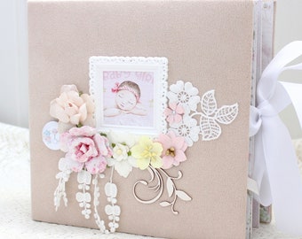 May include: A beige baby photo album decorated with floral embellishments. The album features a framed picture of a sleeping baby, surrounded by pink, white, and yellow paper flowers, lace, and a wooden flourish. A white ribbon ties the album closed.