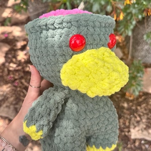 May include: A handmade, plush creature with a green body, a yellow beak, and red eyes. The top of the head is pink. The creature's feet and arms have yellow accents. The toy is made of a soft, textured yarn.
