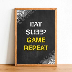 May include: A black chalkboard with a wooden frame, featuring a pixelated design in grey and white. The text "EAT SLEEP GAME REPEAT" is written in white with the word "GAME" highlighted in yellow.
