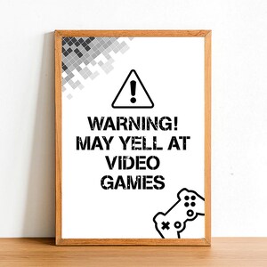 May include: A framed sign with a warning symbol and the text "WARNING! MAY YELL AT VIDEO GAMES". The sign is on a white surface with a wooden frame.