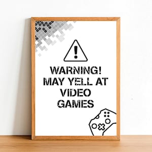May include: A black and white sign with a warning symbol and the text "WARNING! MAY YELL AT VIDEO GAMES". The sign is framed in a light brown wood frame.