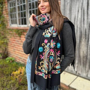 May include: Black scarf with vibrant floral embroidery. The scarf showcases a paisley pattern with pink, blue, and yellow details. It has a fringed edge and is draped around the neck.