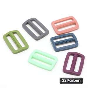 Op de afbeelding: Een verzameling van vijf rechthoekige plastic schuifgespen in olijfgroen, donkergrijs, oudroze, marineblauw, mintgroen en donkerpaars. Elke gesp heeft een centrale balk met verticale ribbels. Een zwart label toont de tekst 22 Farben.