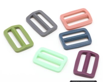 May include: A collection of five rectangular plastic tri-glide sliders in olive green, dark gray, dusty pink, navy blue, mint green, and deep purple. Each slider features a central bar with vertical ridges. A black label displays the text 22 Farben.