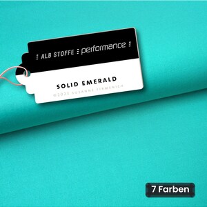 May include: A solid emerald green fabric with a white and black rectangular tag attached by a pink string. The tag features the text ALB STOFFE performance, SOLID EMERALD, and 2023 SUSANNE FIRMENICH. A small black label in the corner reads 7 Farben.