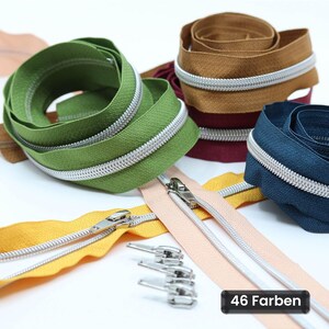 Puede incluir: Una colección de cremalleras enrolladas en varios colores, incluyendo verde, marrón, azul oscuro, amarillo y rosa claro, con dientes y tiradores metálicos plateados. Varios tiradores metálicos plateados sueltos están dispuestos en el centro con el texto 46 Farben.