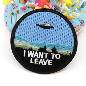 Pode incluir: Um emblema bordado circular apresentando um céu azul claro com um OVNI cinza, uma paisagem verde e cinza com silhuetas de árvores e o texto branco I WANT TO LEAVE costurado em um fundo preto, tudo contornado por uma borda preta.