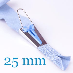 May include: A 25 mm bias tape maker tool constructed from silver-toned metal and light blue plastic. The device features a wire handle and is shown with a strip of light blue fabric being fed through the mechanism to create a folded edge.