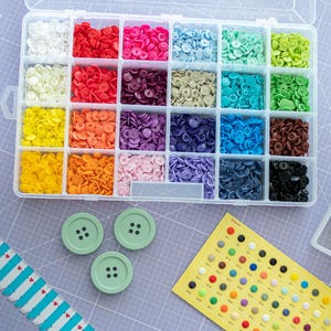 Pode incluir: Um recipiente de armazenamento de plástico transparente com vinte e quatro compartimentos cheios de botões de pressão coloridos em vários tons. Três botões grandes verde-sálvia e uma tabela de referência amarela com pontos coloridos estão próximos.