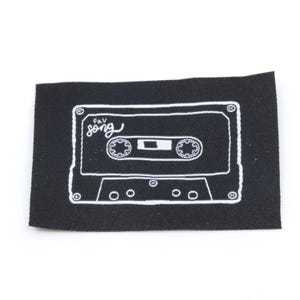 Può includere: Una toppa rettangolare in tessuto nero con il contorno ricamato in bianco di una musicassetta. Il design include il testo "fav song" in corsivo sul lato superiore sinistro del grafico del nastro, con dettagli di bobine e viti.