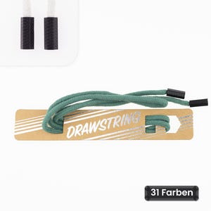 Könnte beinhalten: Eine salbeigrüne Kordel, die um eine rechteckige Pappkarte mit dem in Weiß gedruckten Wort DRAWSTRING gewickelt ist. Die Kordel hat schwarze Kunststoffenden. Ein kleines schwarzes Etikett zeigt den Text 31 Farben.