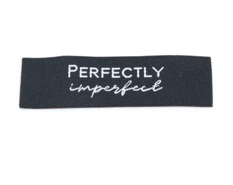 May include: A rectangular black fabric clothing label featuring the word PERFECTLY in a clean, white sans-serif font on the top line, with the word imperfect in a white, cursive, handwritten-style script font directly underneath it.